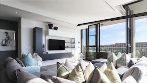 Ein modernes Wohnzimmer mit einem großen grauen Sofa, einem Flachbildfernseher an einer weißen Wand und einem Balkon mit Blick auf die Stadt.