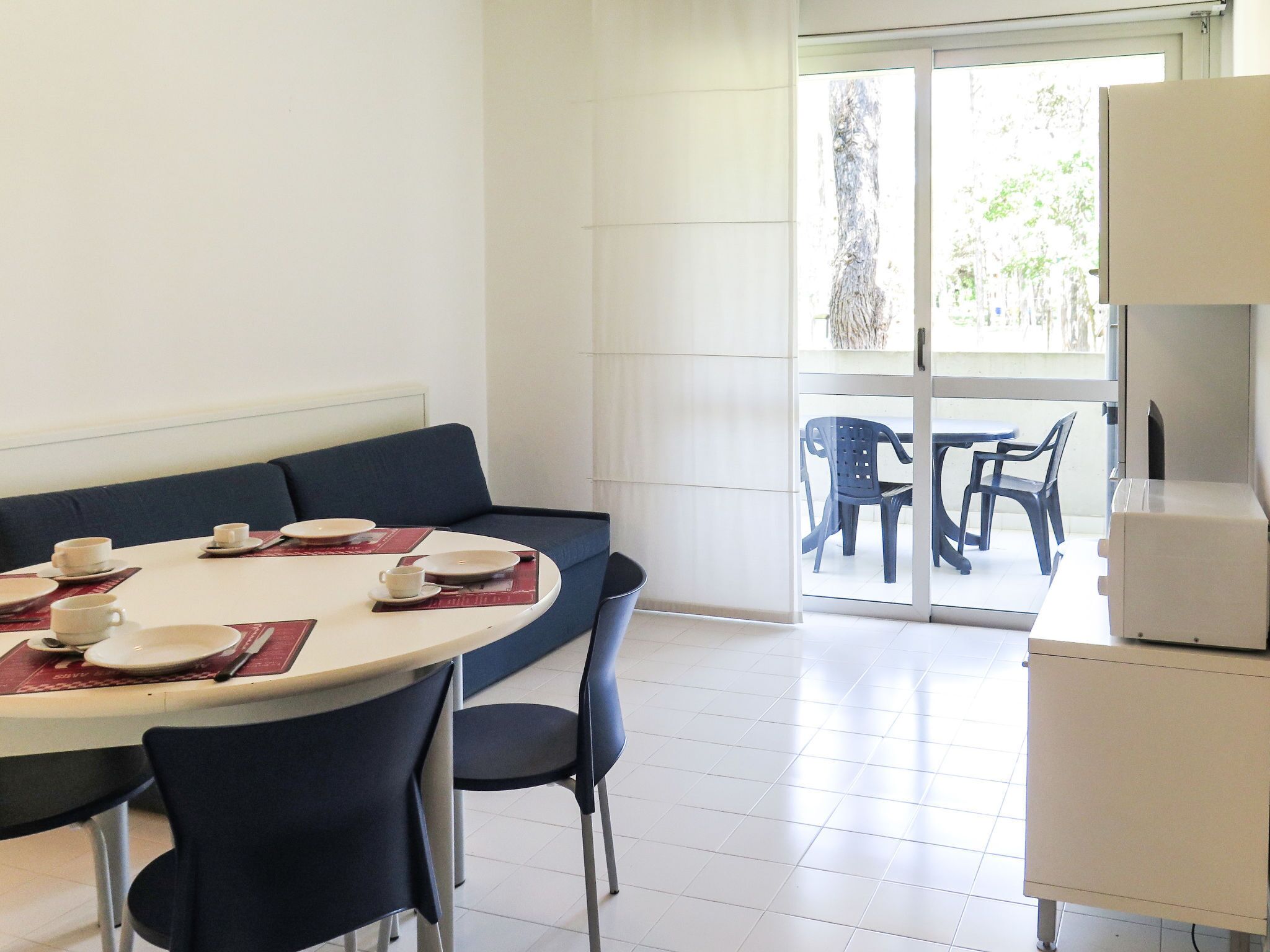 A dining area with a round table, chairs, and a sofa. There is a sliding glass door leading to an outdoor seating area with plastic chairs.