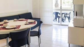 Un comedor con una mesa redonda, sillas y un sofá. Hay una puerta corrediza de vidrio que da a un área de estar exterior con sillas de plástico.