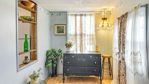 Una habitación con un aparador de madera, un florero verde, una planta en maceta, un estante de madera con objetos decorativos y una ventana con cortinas.