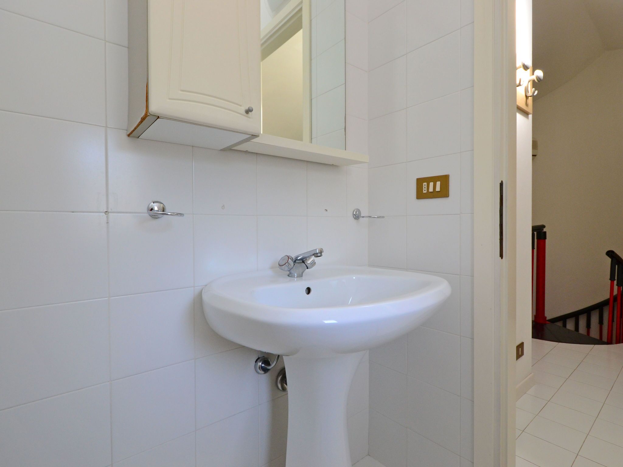 Un bany blanc amb un lavabo amb peu, un mirall a sobre i una porta que condueix a una altra habitació.