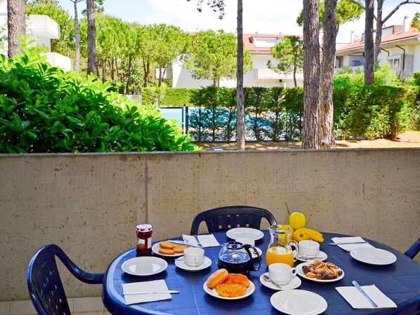 A round table set for breakfast with plates, cups, and a pitcher of orange juice, surrounded by greenery and trees.