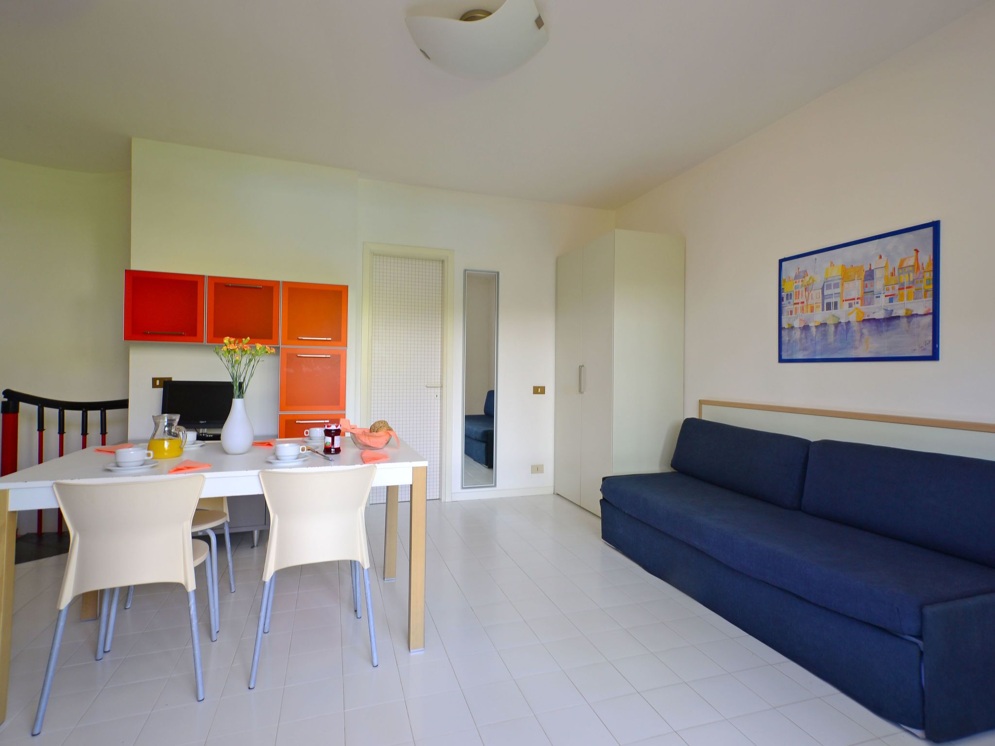 A modern living room with a blue sofa, a dining area with a white table and orange cabinets, and a painting on the wall.