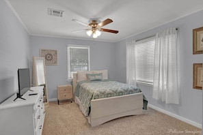 A bedroom with a bed, a desk with a computer, a ceiling fan, and two windows with blinds.
