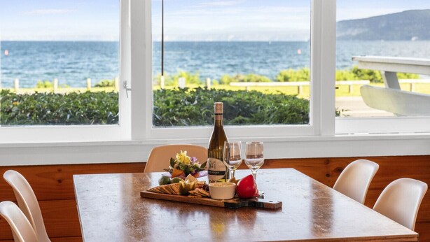 A dining table with a bottle of wine, glasses, and a fruit platter by a window overlooking the sea. - 3 Oaks on the Lake -  Kinloch (Kinloch)