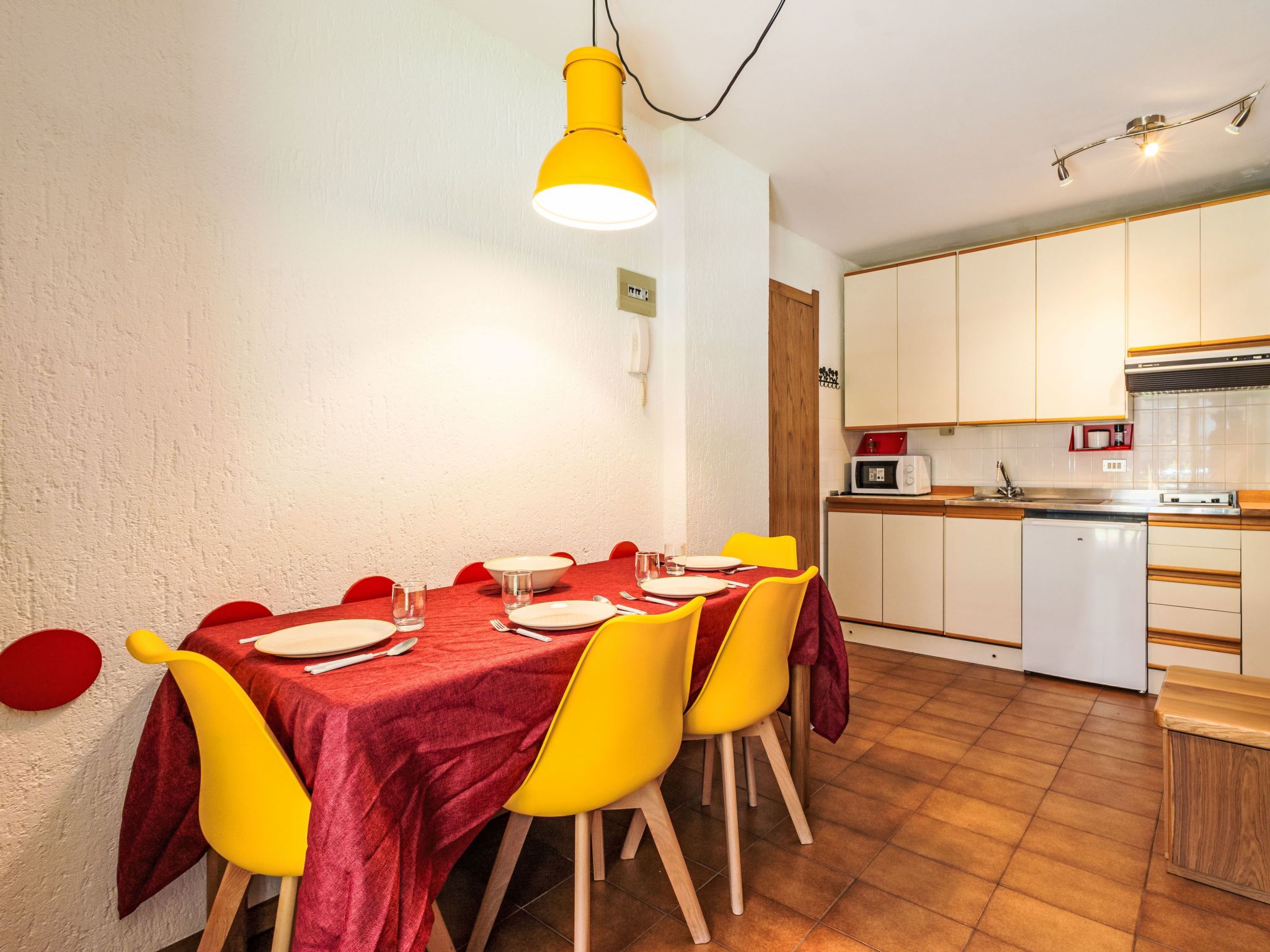 Een eetgedeelte met een tafel voor vier personen, gele stoelen en een rode tafelkleed. Er is een keuken met witte kasten en een magnetron.