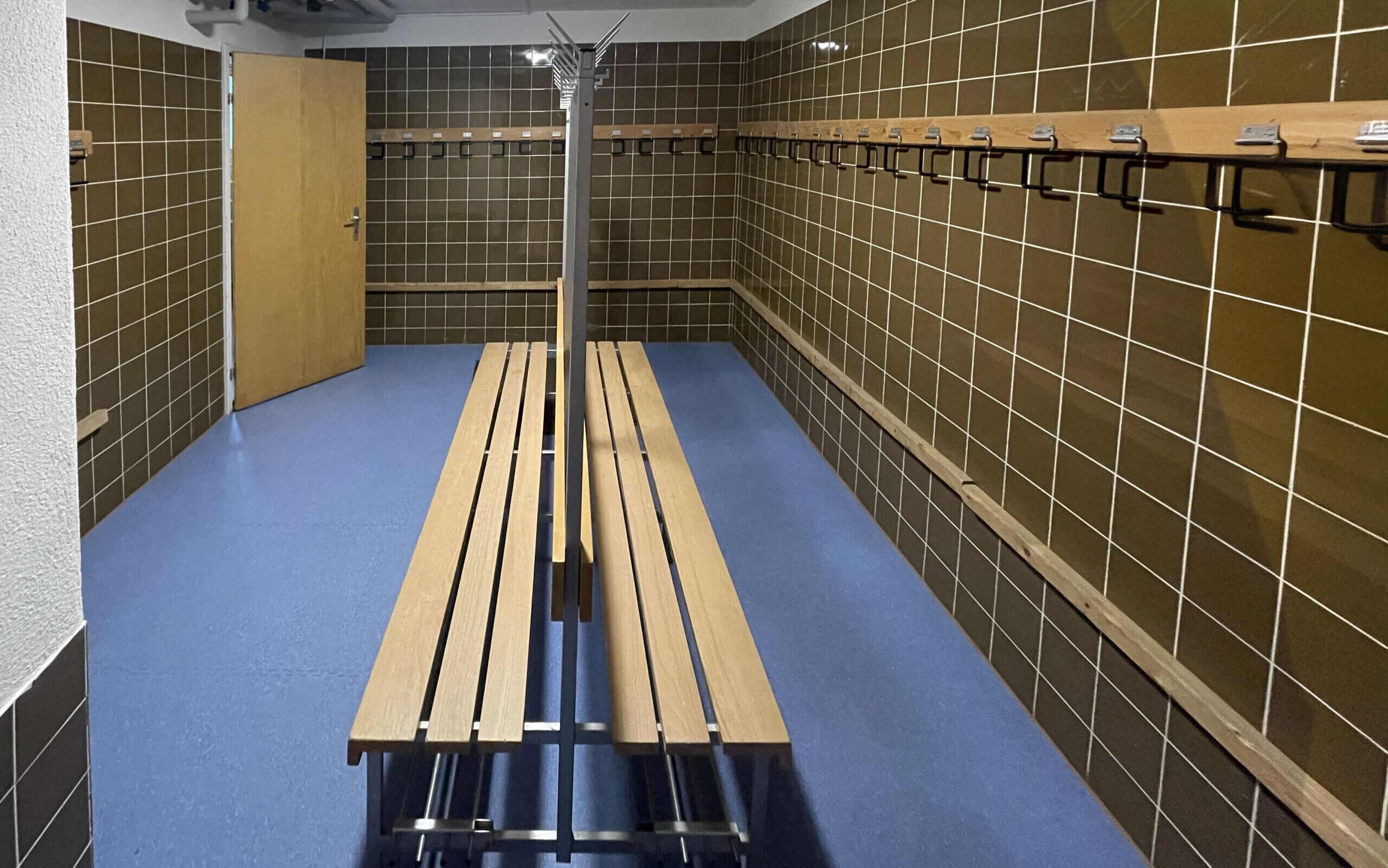 Un vestuario con suelo azul, bancos de madera y taquillas metálicas en las paredes.