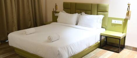 A neatly made bed with white linens and rolled towels, a green headboard, and a nightstand with a lamp.
