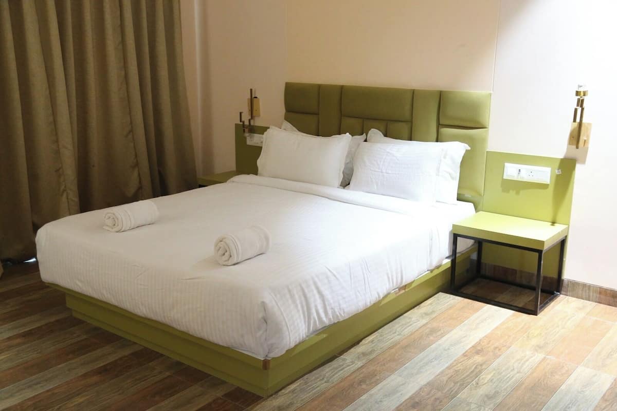 Una cama bien hecha con ropa blanca y toallas enrolladas, un cabecero tapizado en verde, una mesita de noche con lámpara y suelo de madera.