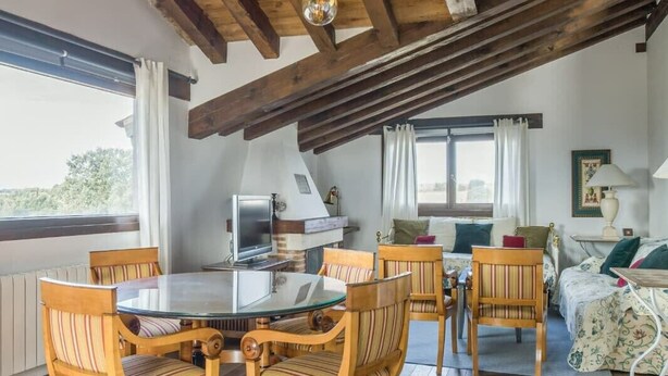A cozy dining area with a round glass table surrounded by wooden chairs with striped cushions. A wooden ceiling with exposed beams and a hanging light fixture is visible above. - Finca Montechico, Casa Matute, rural Segovia (Segovia)