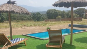 A pool area with two wooden lounge chairs and thatched umbrellas, surrounded by green artificial grass and a view of distant trees and mountains. - Finca Montechico, Casa Matute, rural Segovia (Segovia)