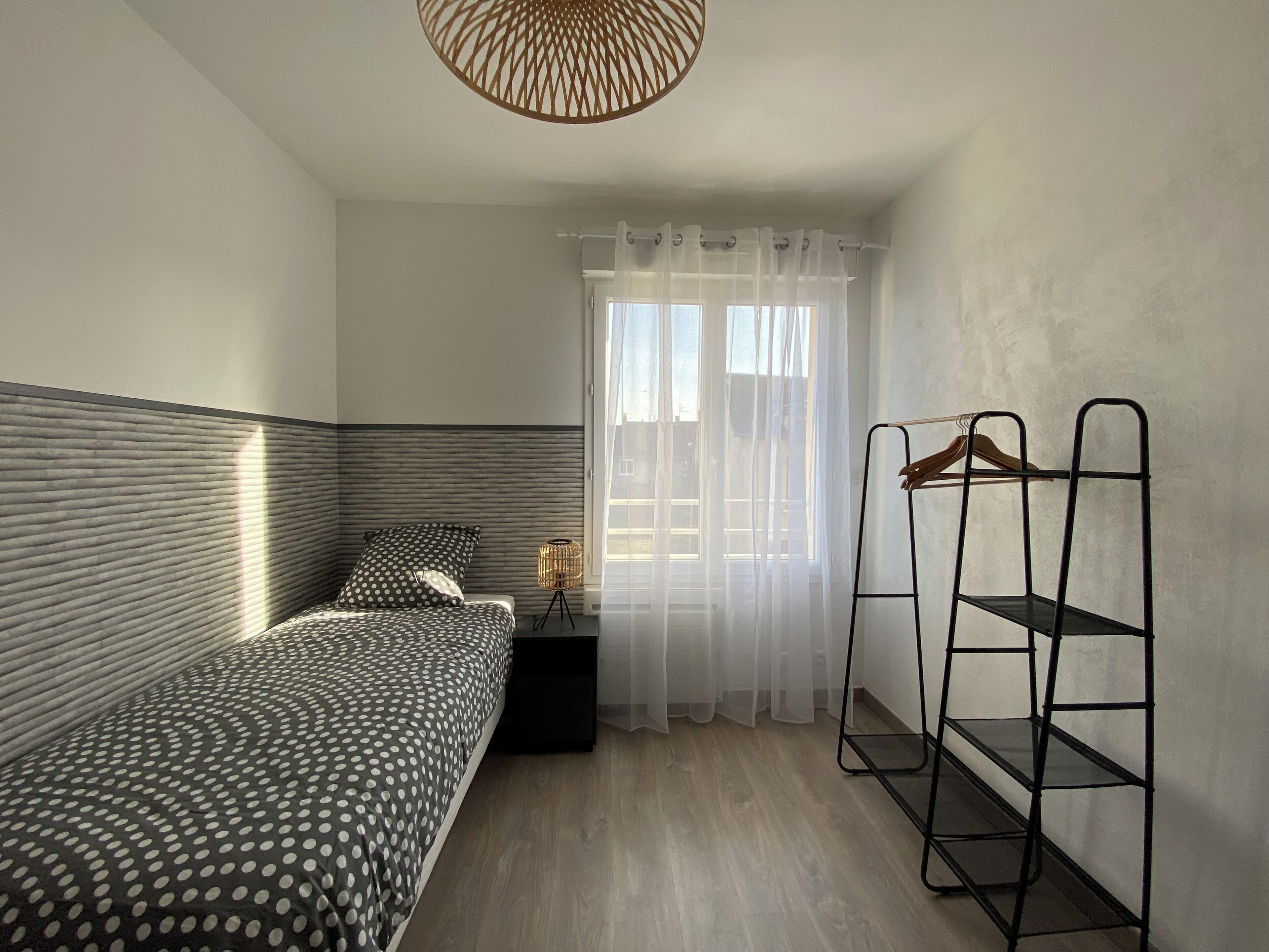 A bedroom with a bed, a bedside table, a hanging rack, and a window with sheer curtains.