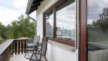 A balcony with a table and chairs, a window with a view of trees and buildings, and a metal railing.