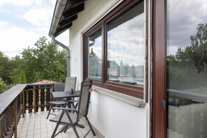 Een balkon met een tafel en stoelen, een raam met uitzicht op bomen en gebouwen, en een metalen hekwerk.