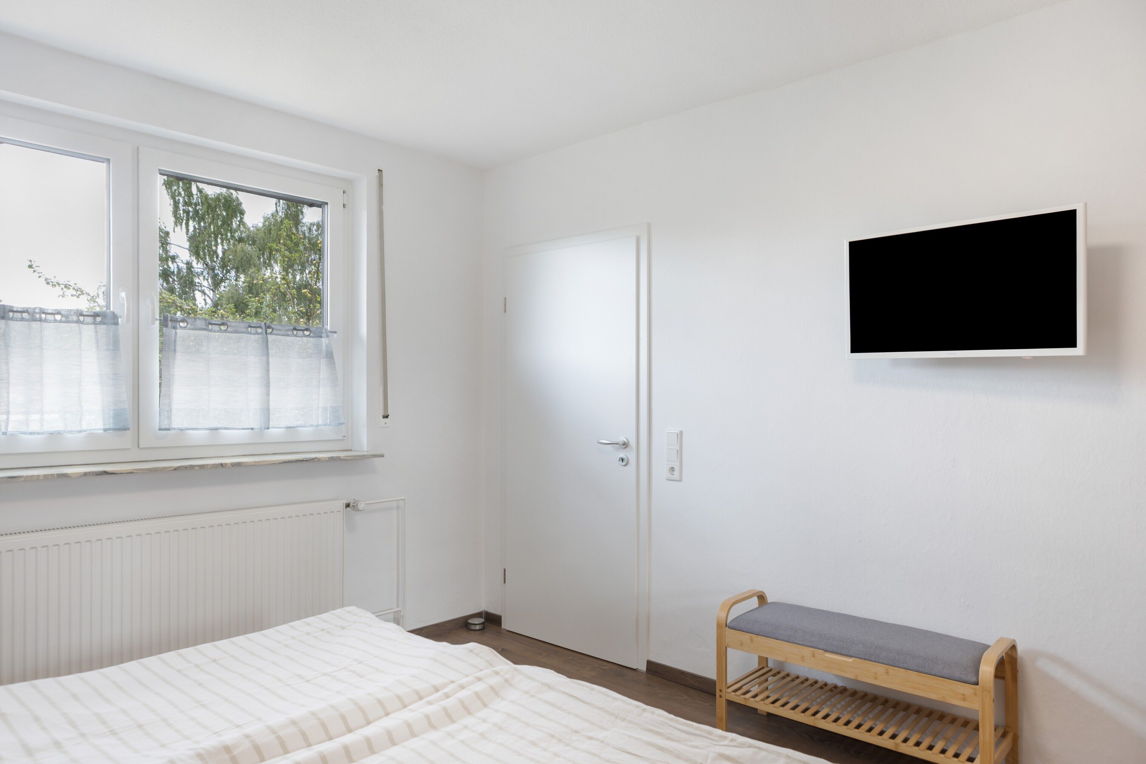 Ett sovrum med en säng, en bänk, en tv, en dörr och ett fönster.