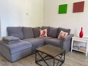 Ein graues Ecksofa mit einem Holztisch, ein weißer Beistelltisch mit einer roten Vase und ein Bücherregal mit Zeitschriften.