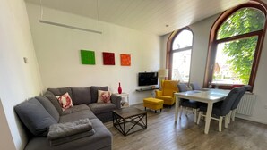 A living room with a grey sofa, a yellow armchair, a dining table with chairs, a TV, and a window with a view of greenery. - Apartment 'Nickel 1' with Shared Garden and Wi-Fi (Arnis)
