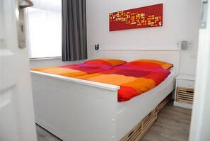 Ein weißes Bett mit bunter Bettwäsche, ein abstraktes rotes Bild an der Wand und ein Fenster mit Vorhängen.