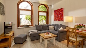 Ein Wohnzimmer mit grauem Sofa, einem Holztisch, einem Fernseher auf einem Schrank und einem Fenster mit Blick auf ein Segelboot.