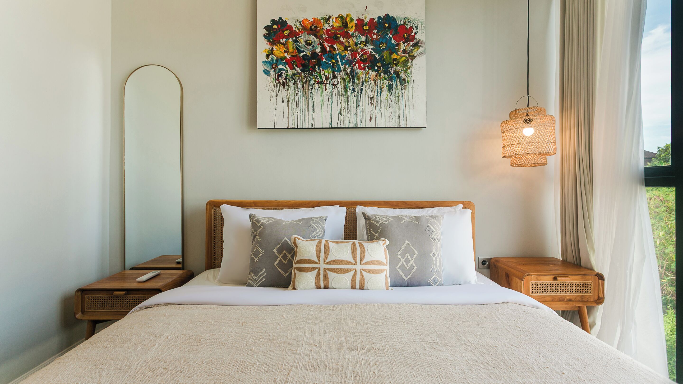 A neatly made bed with a patterned throw, a large abstract painting above it, and a hanging light fixture.