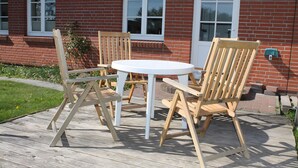 A brick building with a white door and windows, a wooden deck with a round table and four chairs, and a small plant near the door. - Holiday Home 'Ferienhaus Stie Tinningstedt' with Private Terrace, Private Garden and Wi-Fi (Tinningstedt)