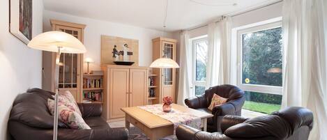 Un salon comprenant un canapé, un fauteuil, une table, une lampe, une bibliothèque et une fenêtre avec des rideaux.