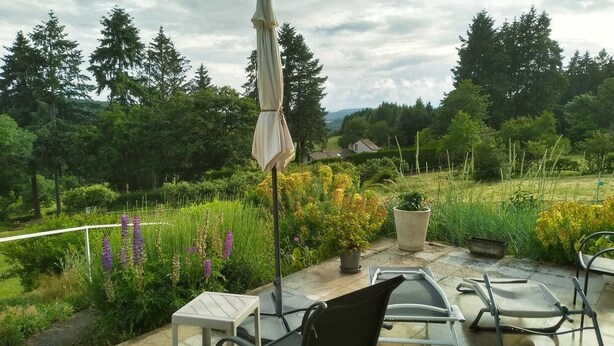 An outdoor patio with a table and chairs, surrounded by lush greenery and a view of trees and hills. - Gîte at the gateway to the Morvan (Magnien)