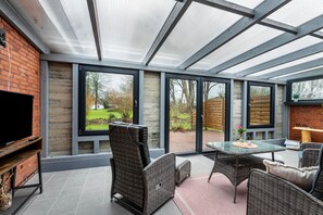 Een moderne patio met rieten meubilair, een glazen tafel en een televisie aan een bakstenen muur.