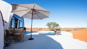 A desert landscape with a pool, outdoor furniture, and a large umbrella. - Reverie Kalahari Pod (Mariental)