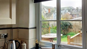 Una cocina con un lavamanos de acero inoxidable doble, un hervidor y una ventana con vistas a un jardín y casas.