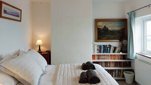 Una cama bien hecha con almohadas, una lámpara de noche, una estantería con libros, un cuadro y una ventana con cortinas.