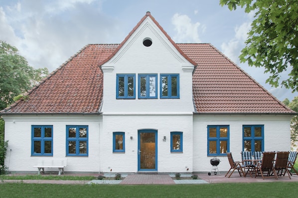Une maison blanche à deux étages, avec un toit recouvert de tuiles rouges, un porche à l’avant doté d’un banc et un barbecue.