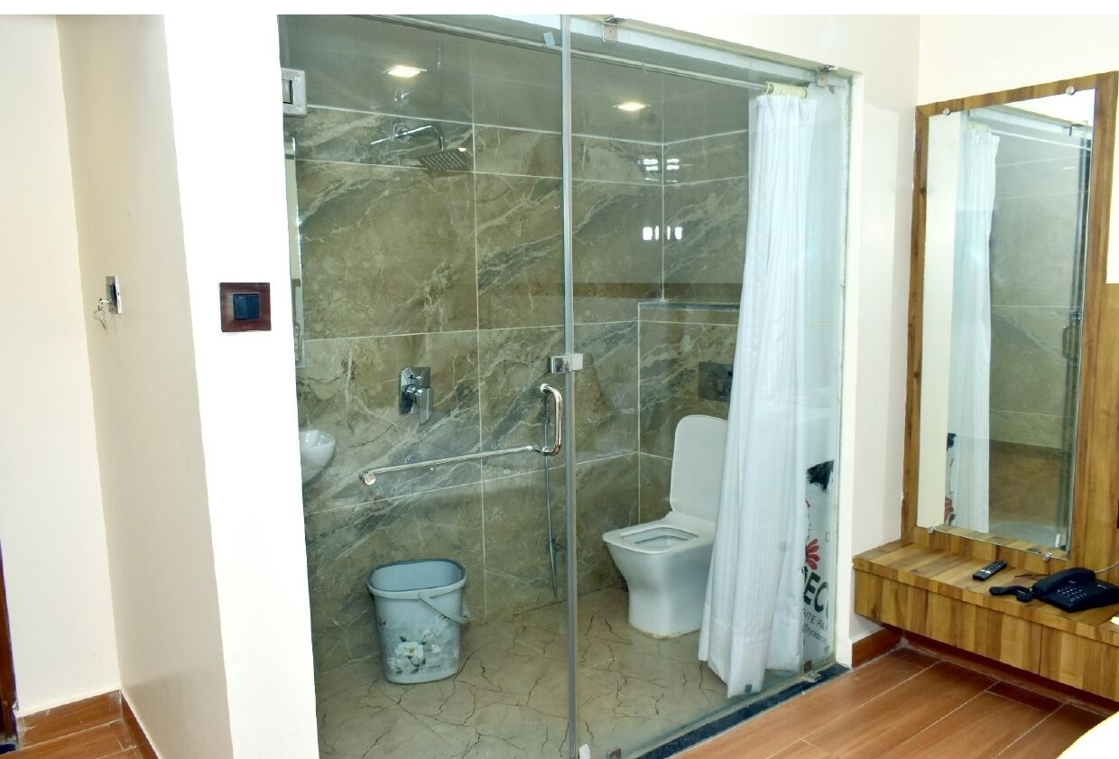 A modern bathroom with a glass-enclosed shower, a toilet, a small trash bin, and a wooden bench with a mirror and phone.