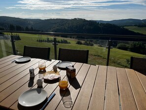 A wooden table set with plates, cutlery, a glass of orange juice, and a box of cereal on a balcony with a scenic view of hills and fields. - Holiday Home 'Winterberg D4' with Mountain View, Shared Terrace and Wi-Fi (Winterberg)