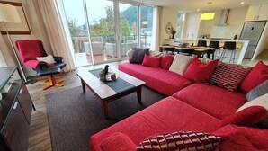 A modern living room with a red sofa, a wooden coffee table, and a red armchair. There is a balcony with a view of greenery and mountains. - Stunning Views, 2 Bedroom 2 minutes to town centre (Queenstown)