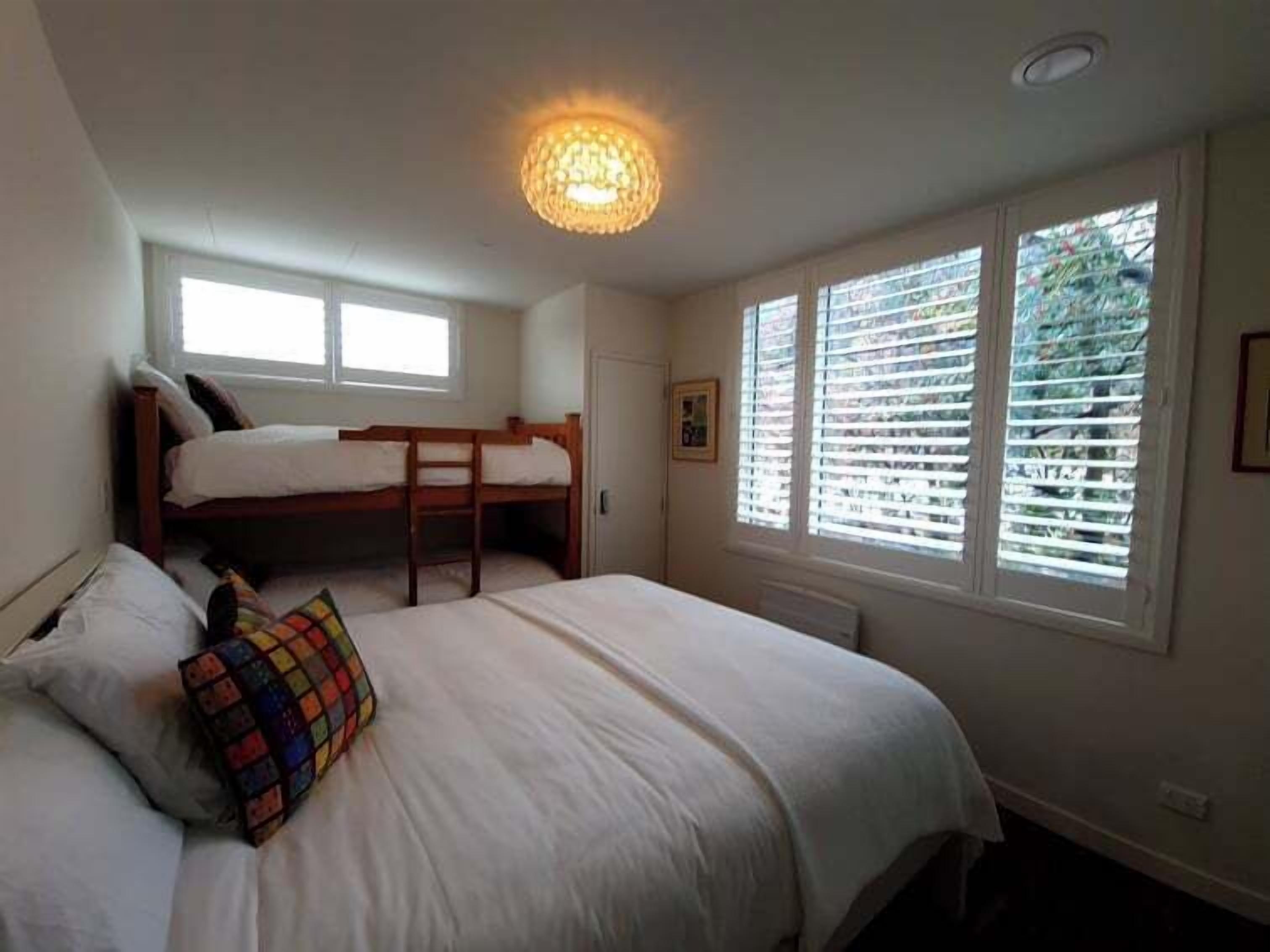 A bedroom with a bunk bed, a single bed, a window with blinds, and a ceiling light fixture.