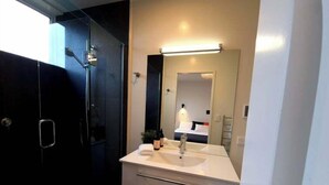A modern bathroom with a glass shower enclosure, a white sink with a mirror above it, and a small bedside table with a bottle and a plant. - Stunning Views, 2 Bedroom 2 minutes to town centre (Queenstown)