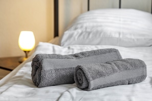 A neatly made bed with white linens and a rolled gray towel.