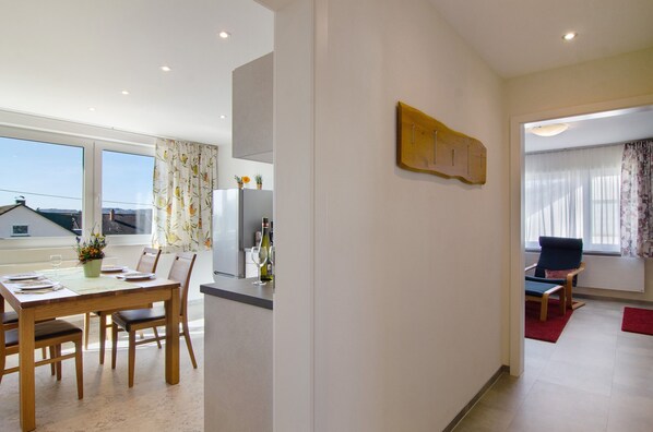 A modern dining area with a wooden table and chairs, a kitchen in the background, and a view through the window.