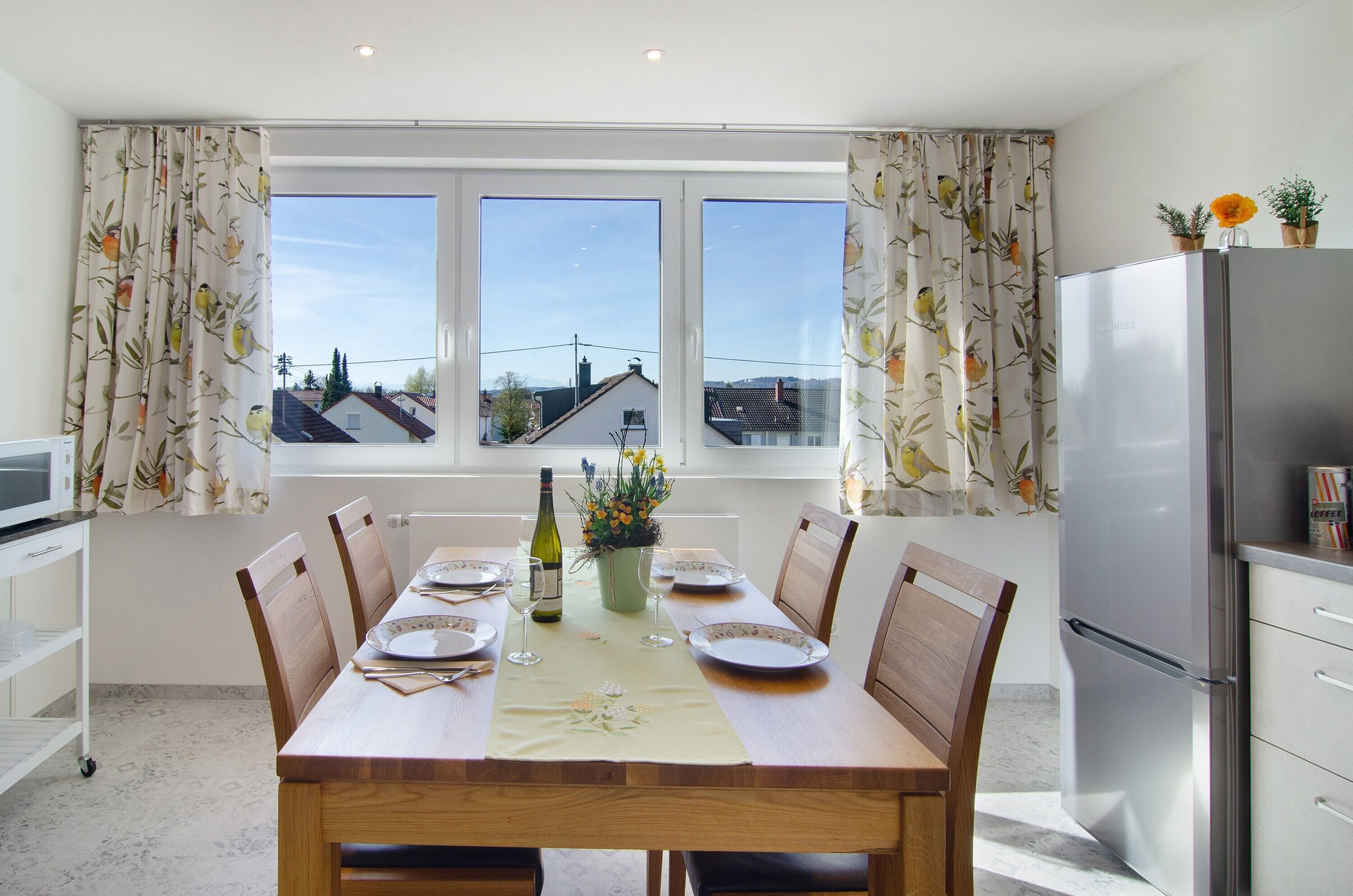 A dining area with a table set for six, a refrigerator, and a window with curtains.