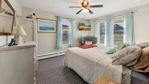 A bedroom with a bed, a ceiling fan, a desk with a lamp, a chair, and a window with curtains.