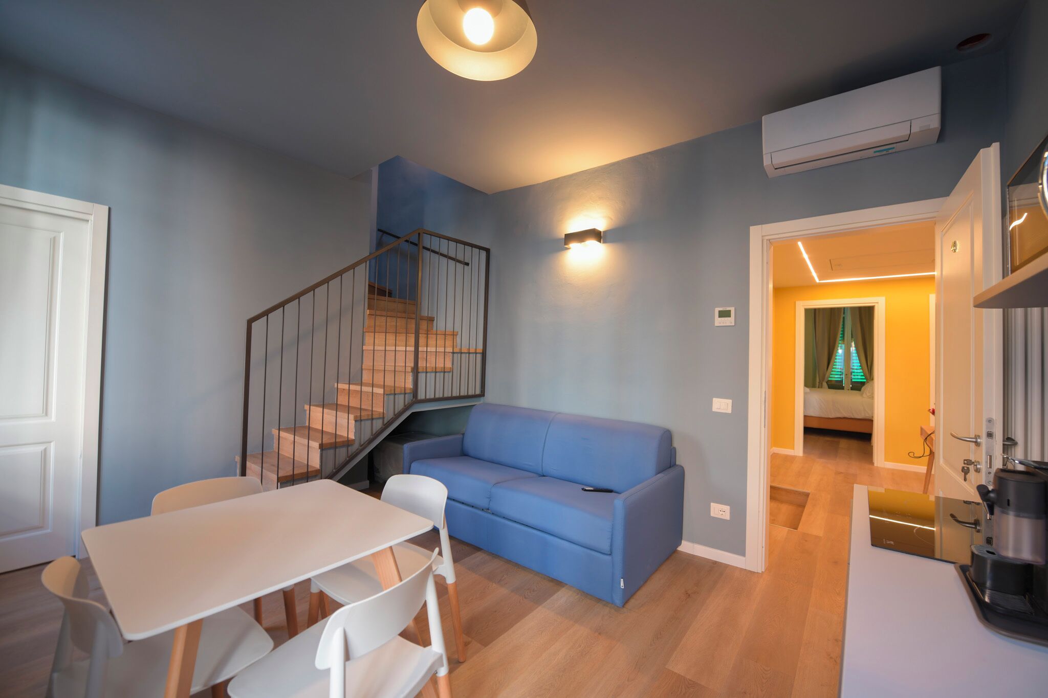 A compact living space with a staircase, a sofa, a dining table, and a kitchen area.