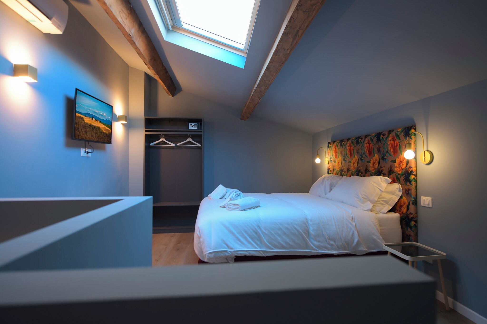 A bedroom with a skylight, a bed with white linens, a wall-mounted TV, and a floral-patterned headboard.
