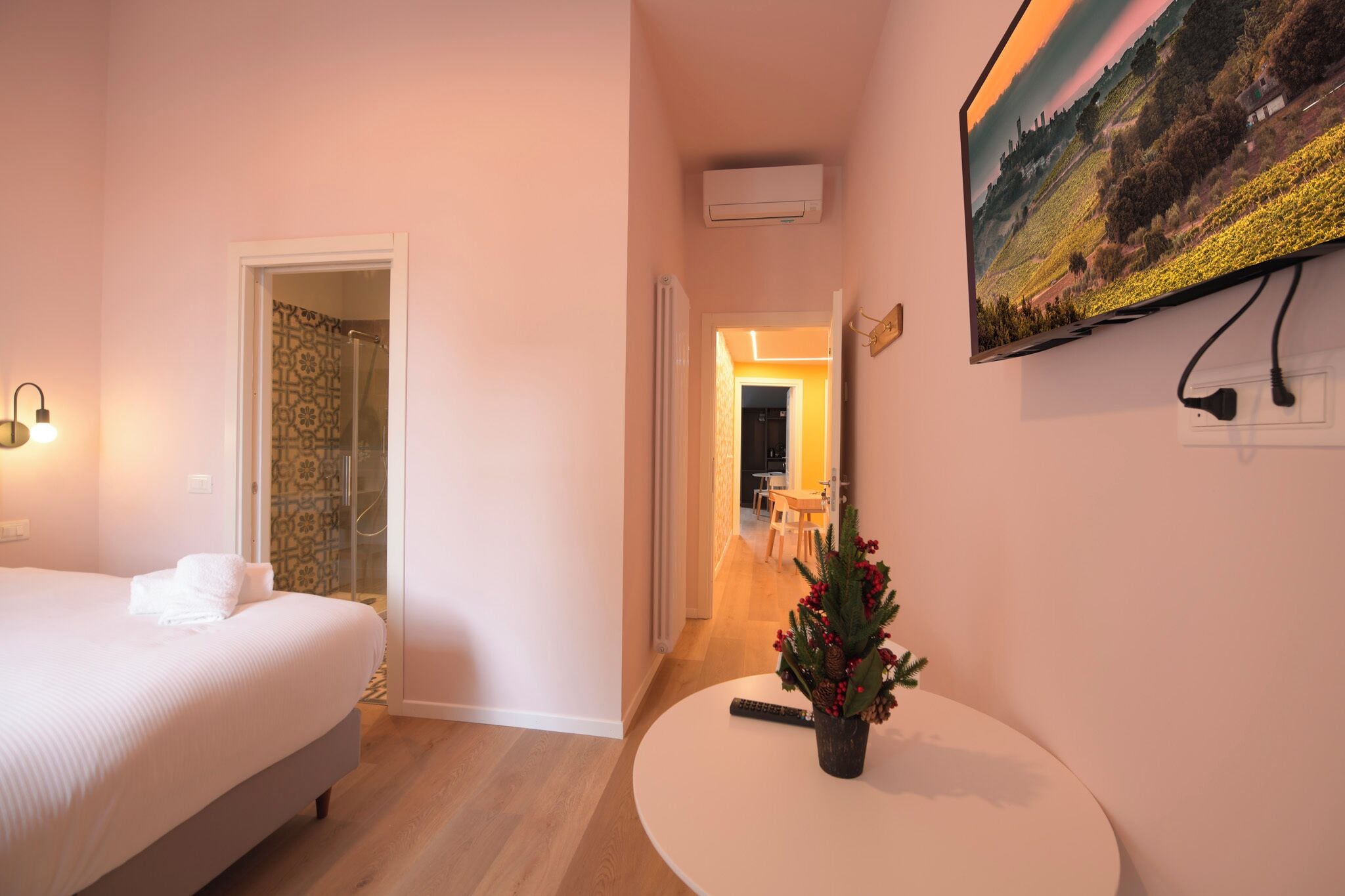 Una camera d'albergo con un letto, un tavolino con una pianta, un quadro appeso alla parete e un corridoio che porta a un'altra stanza.