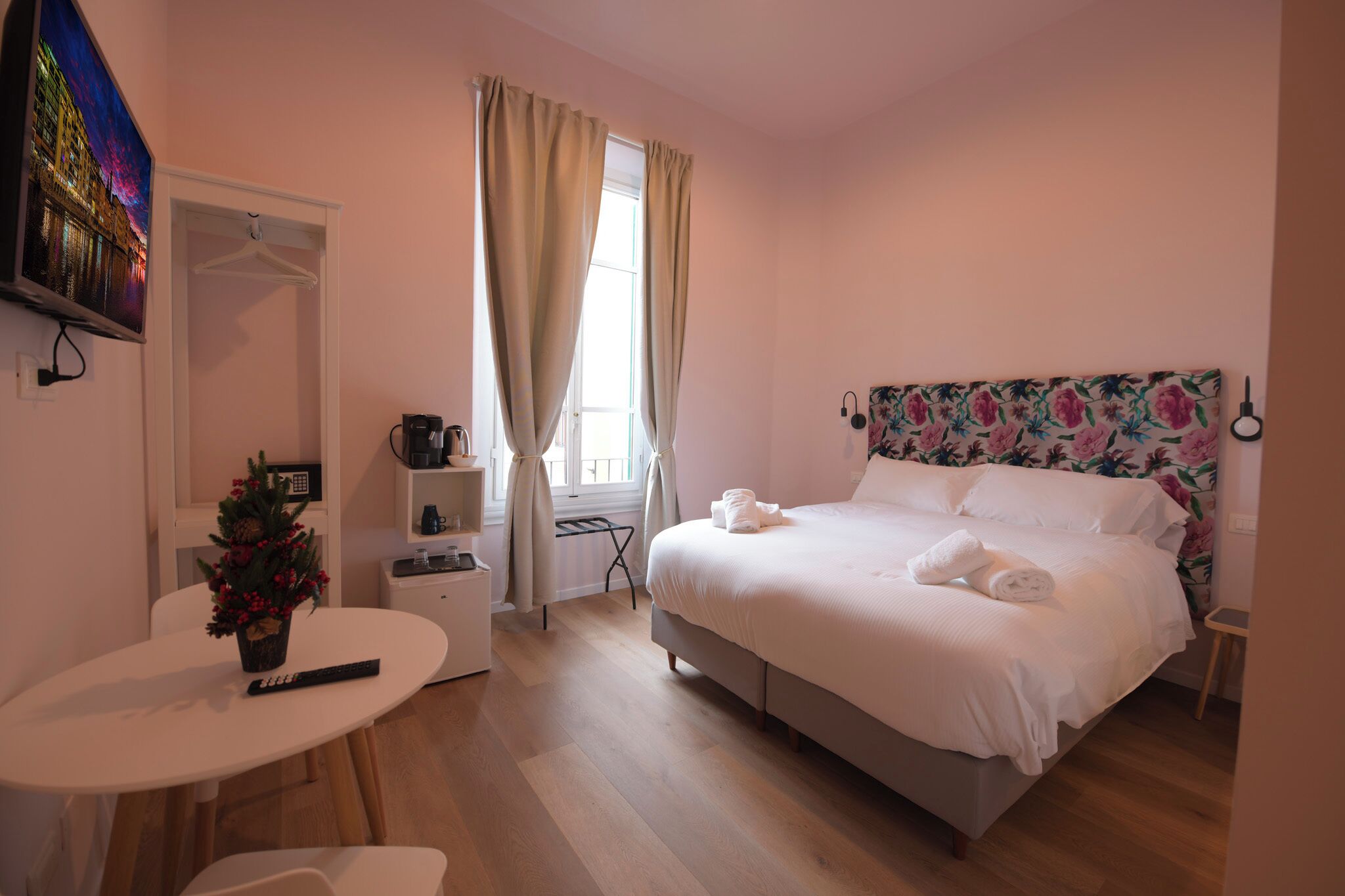 Une chambre à coucher comprenant un lit, une petite table, une chaise, un téléviseur, une fenêtre avec des rideaux et une œuvre d’art accrochée au mur.