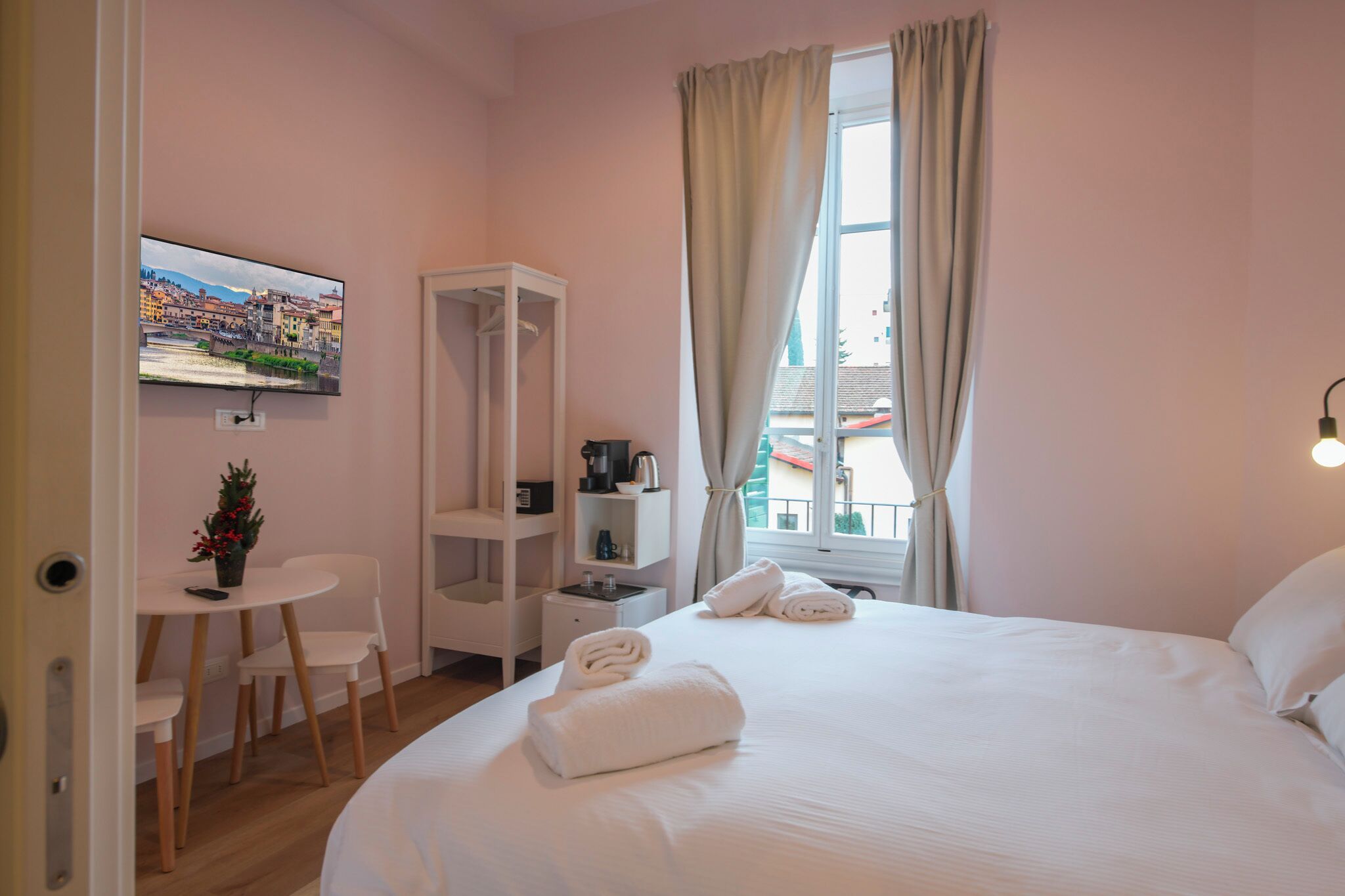 A hotel room with a bed, a TV mounted on the wall, a small table with chairs, a wardrobe, and a window with curtains.