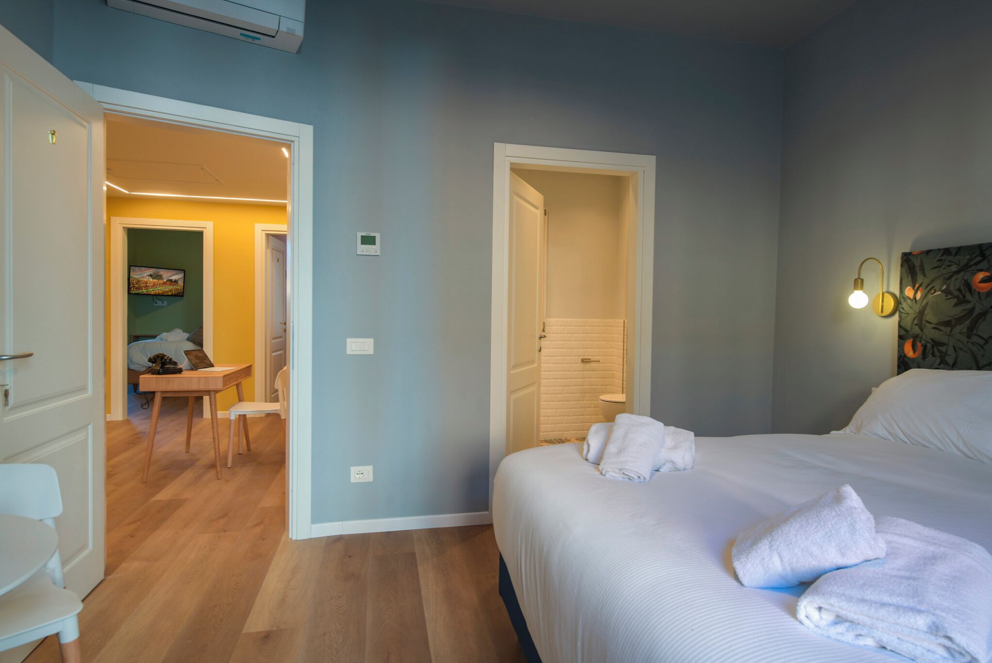 A hotel room with a bed, a desk, and a chair. There is a door leading to another room.