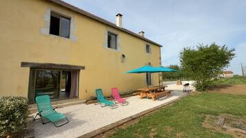 A yellow building with a patio area featuring a wooden table, four lounge chairs, and a blue umbrella.