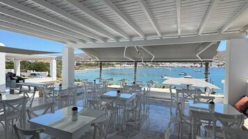 Un área de estar al aire libre con mesas y sillas blancas, con vistas a un puerto deportivo con barcos y un mar azul cristalino.