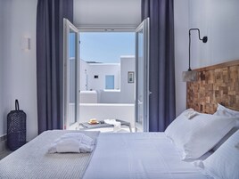 Habitación de hotel con cama, una maleta, una mesa con un sombrero y gafas de sol, y vista a un edificio blanco a través de una puerta abierta.